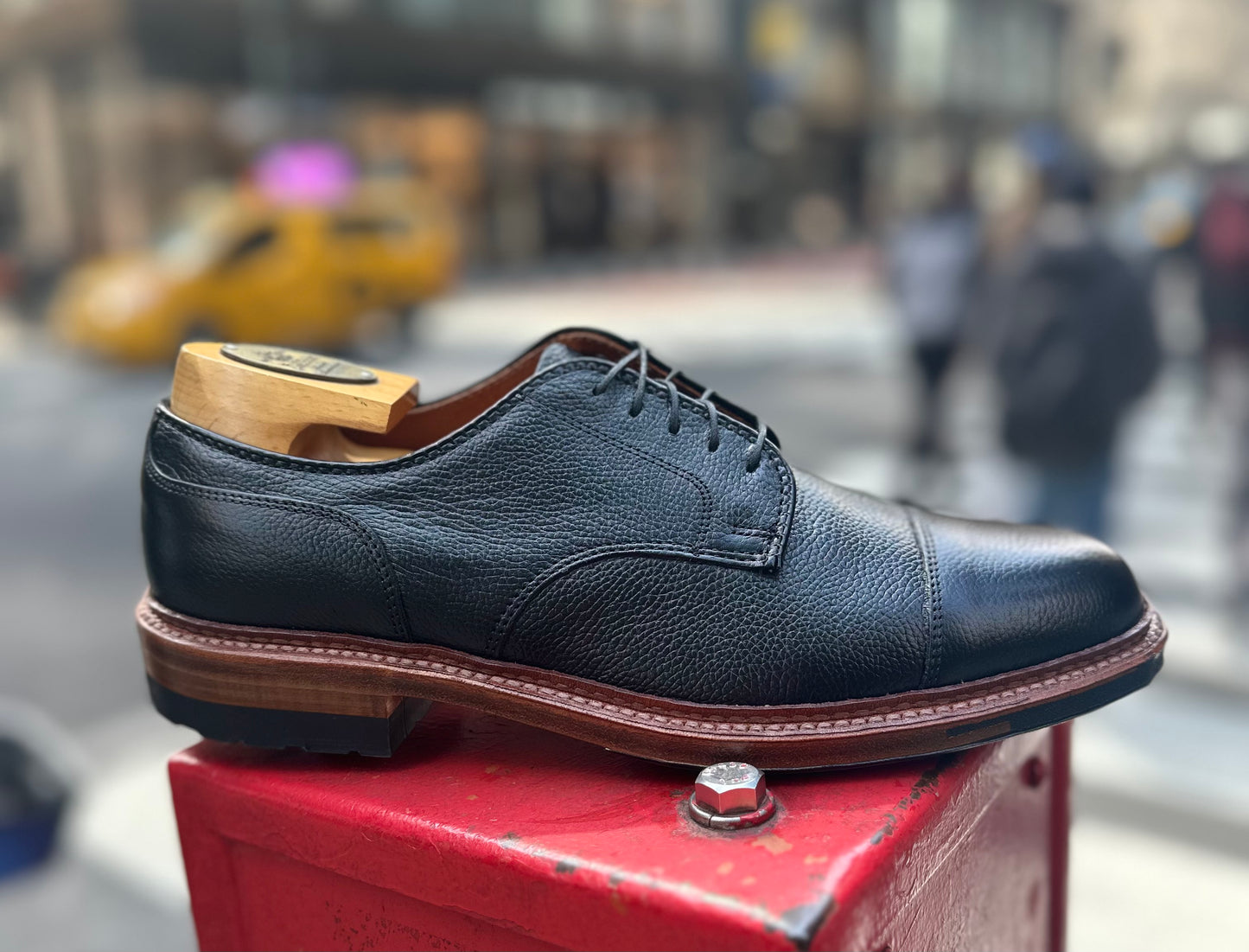 The Alden Madison 22826C - Black JS Grain "Derosa" Blucher, paired with a wooden shoe tree, is shown outdoors on a red metal surface with a blurred city street and people in the background.