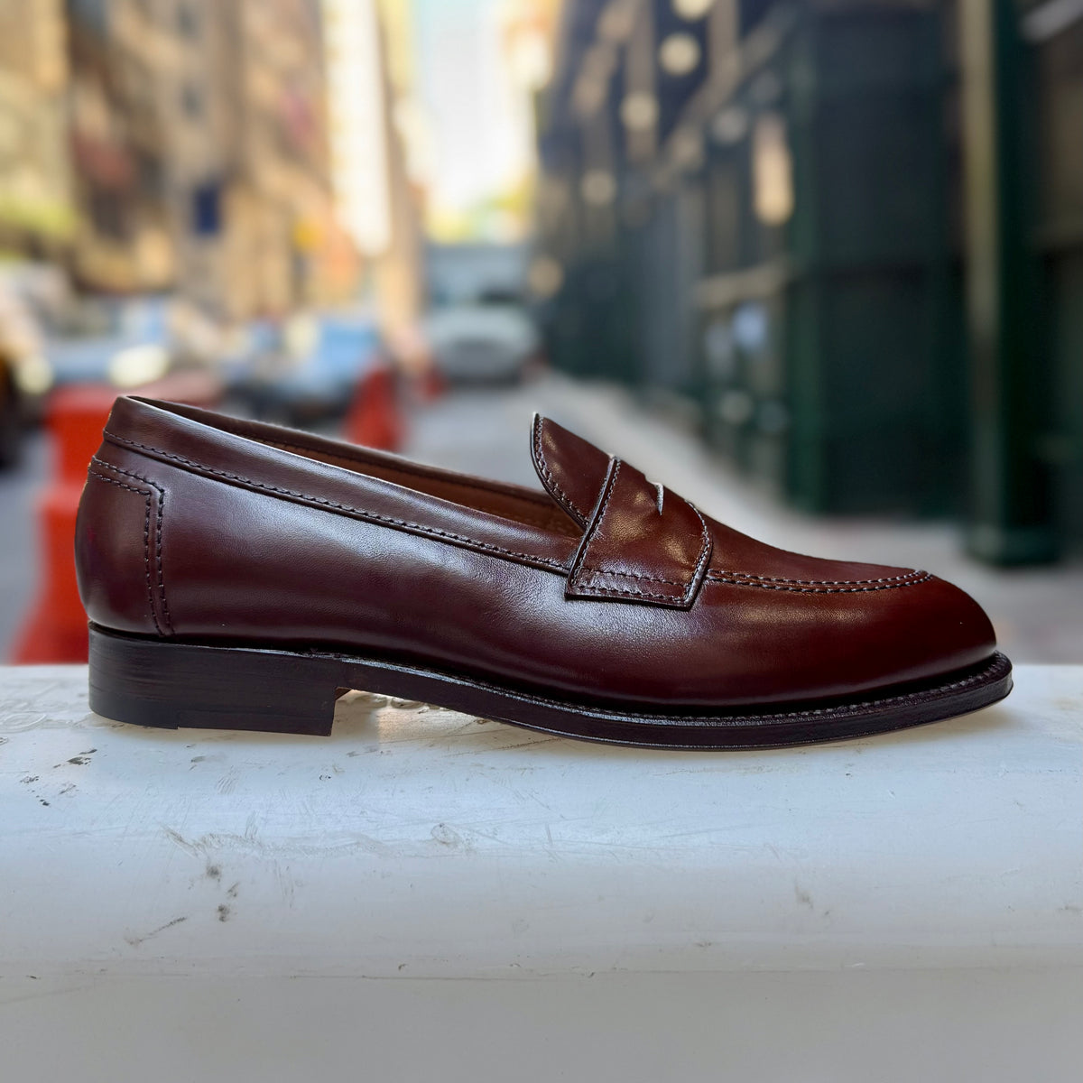 The D5214 - Brown Calf Flex Penny by Alden Madison, crafted from Brown Vegano Calf leather on the refined Copley Last with a Flex Leather Sole, is displayed on a white surface with a blurred city street scene in the background.