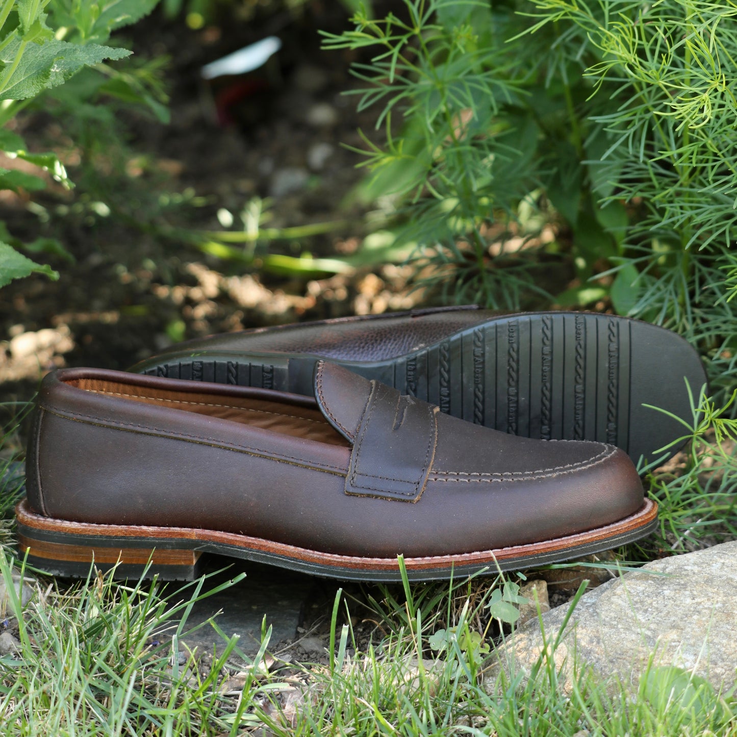 The ALDEN SHOES 5730L- Penny Mocc in Kudu with rubber sole is shown outdoors on grass near green plants and rocks, featuring visible stitching, a classic mocc toe, and brown kudu leather.