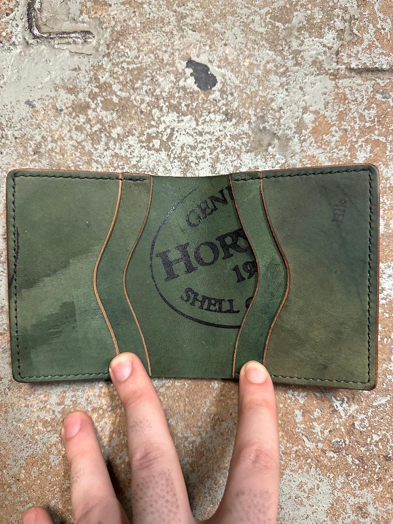 A hand displays the Alden Madison Herb Bi-Fold Wallet in Shell Cordovan, featuring green leather, brown stitching, wavy card slots, and a visible Horween Shell Cordovan stamp against a textured background.