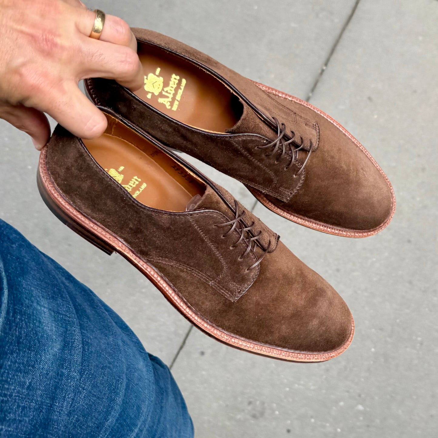 A person holds a pair of Alden Madison 29335F - Unlined Dover dress shoes in brown suede by the heels, standing on a gray sidewalk and wearing blue jeans.