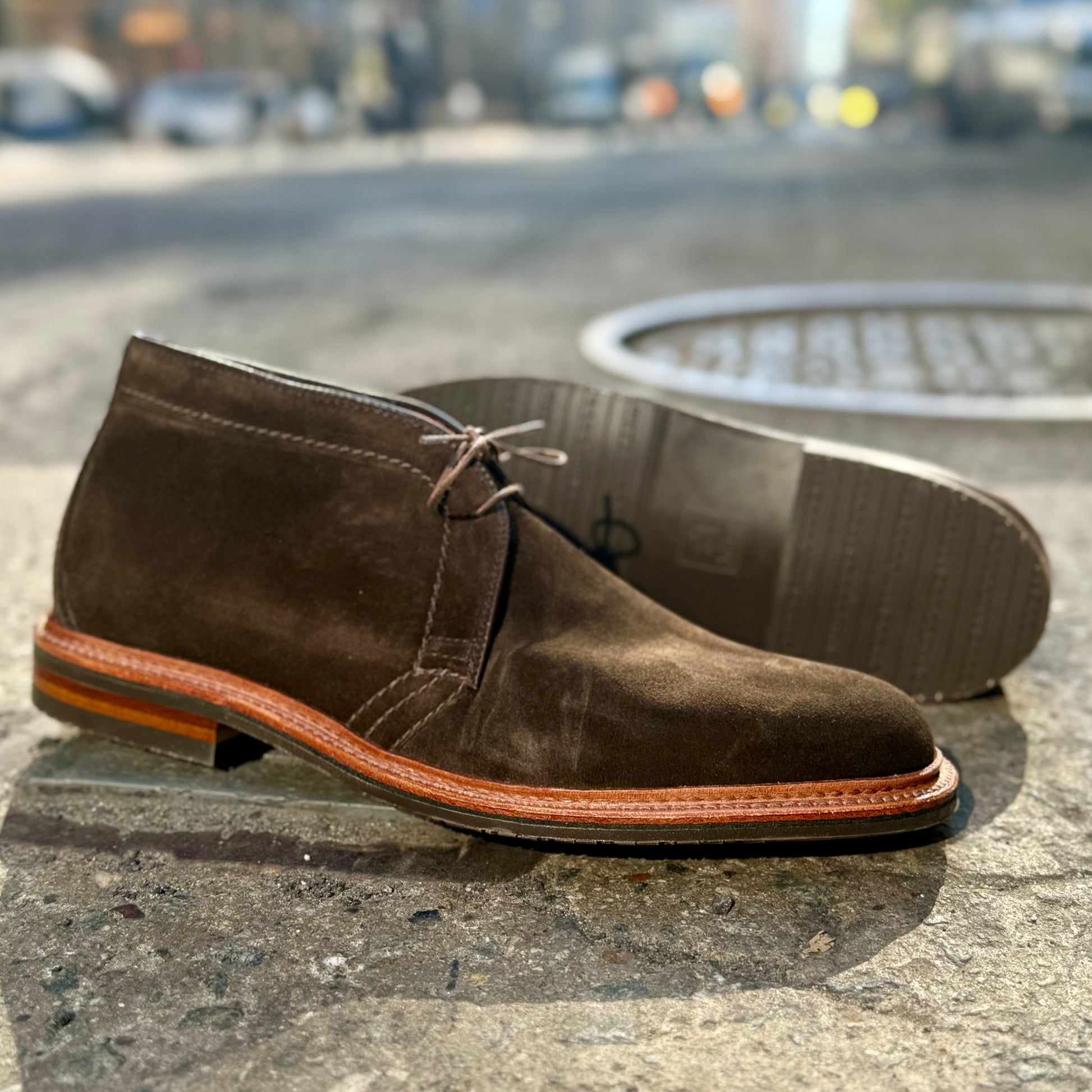 The D1712L Chukka Boot by Alden Madison, crafted on the Barrie Last in Loden Suede with a reverse welt, features a tan sole and laces on a sunlit sidewalk. Next to it, another boot lies on its side, highlighting its unique tread against a blurred urban backdrop.
