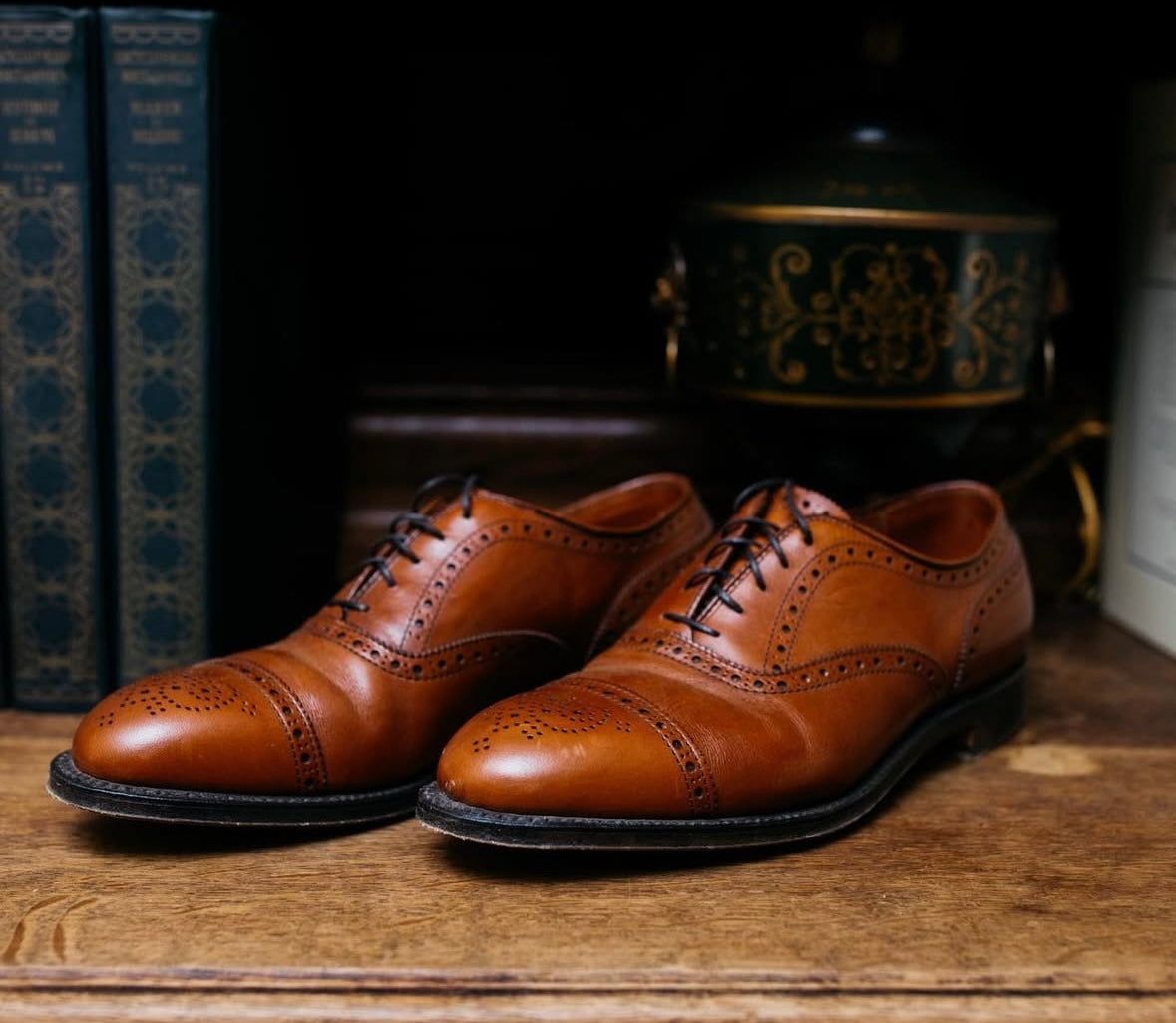 The Alden Madison 911 - Burnished Tan Medallion Cap Toe Bal dress shoes, featuring calfskin leather and brogue details, rest on a wooden surface between vintage books and a decorative container.