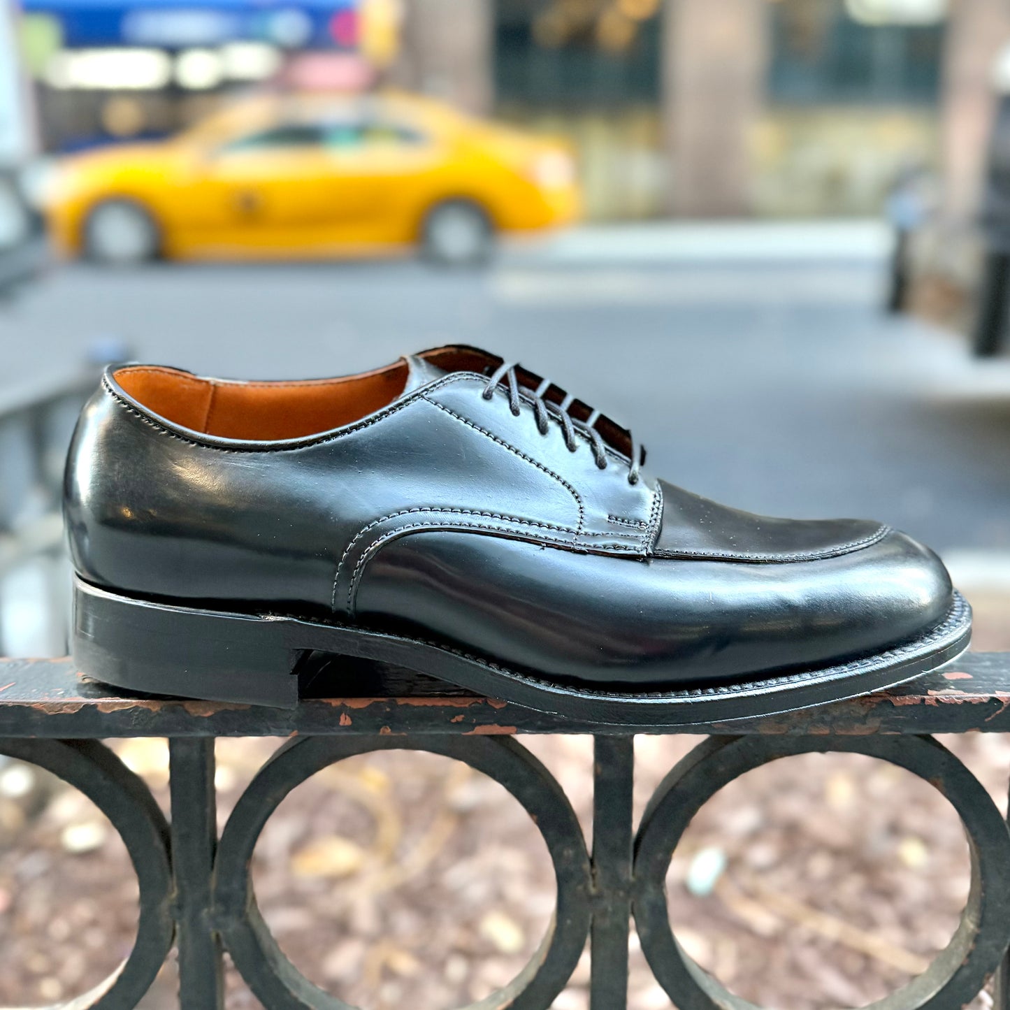 The Alden Madison N6406 - Black Shell V-Tip on 379X is shown outdoors on a metal railing with laces, Military Last, and Rubber Heel, against a blurred city street and yellow taxi backdrop.