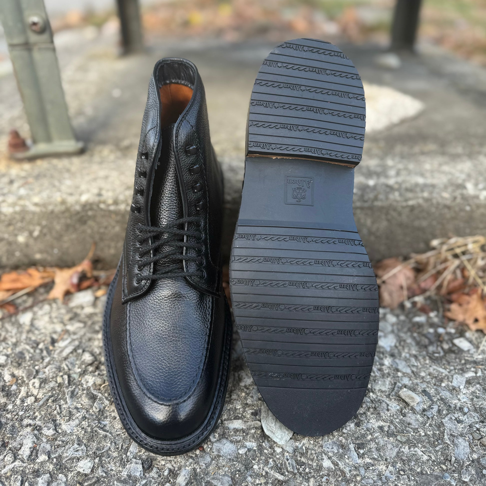 A pair of Alden Madison D6956LH "Han" Boots in Black JS Grain, one upright and one on its side to show the textured rubber sole, rest on rough concrete scattered with dry leaves.