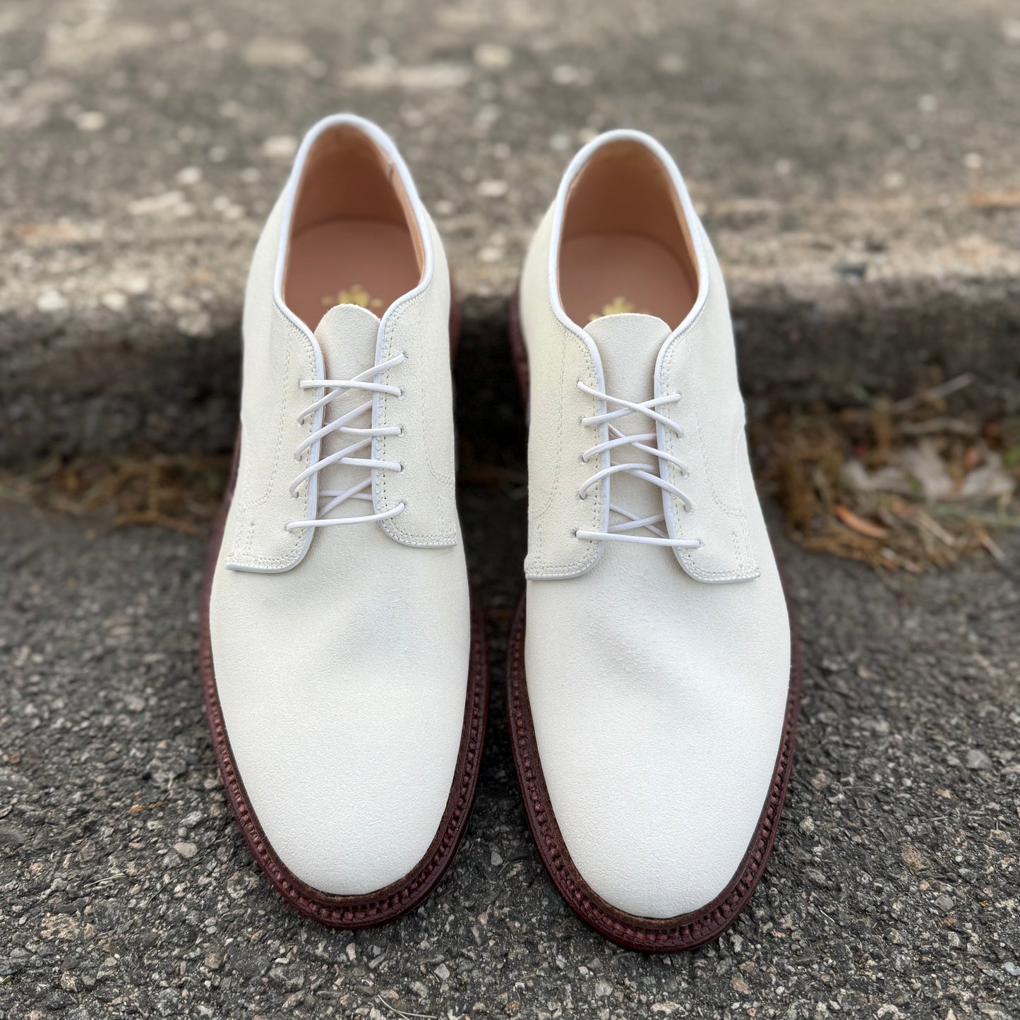 The Alden Madison 29330F Unlined Dover in ivory suede, featuring classic lace-up styling and brown leather soles, is displayed side by side on textured pavement near a concrete step.