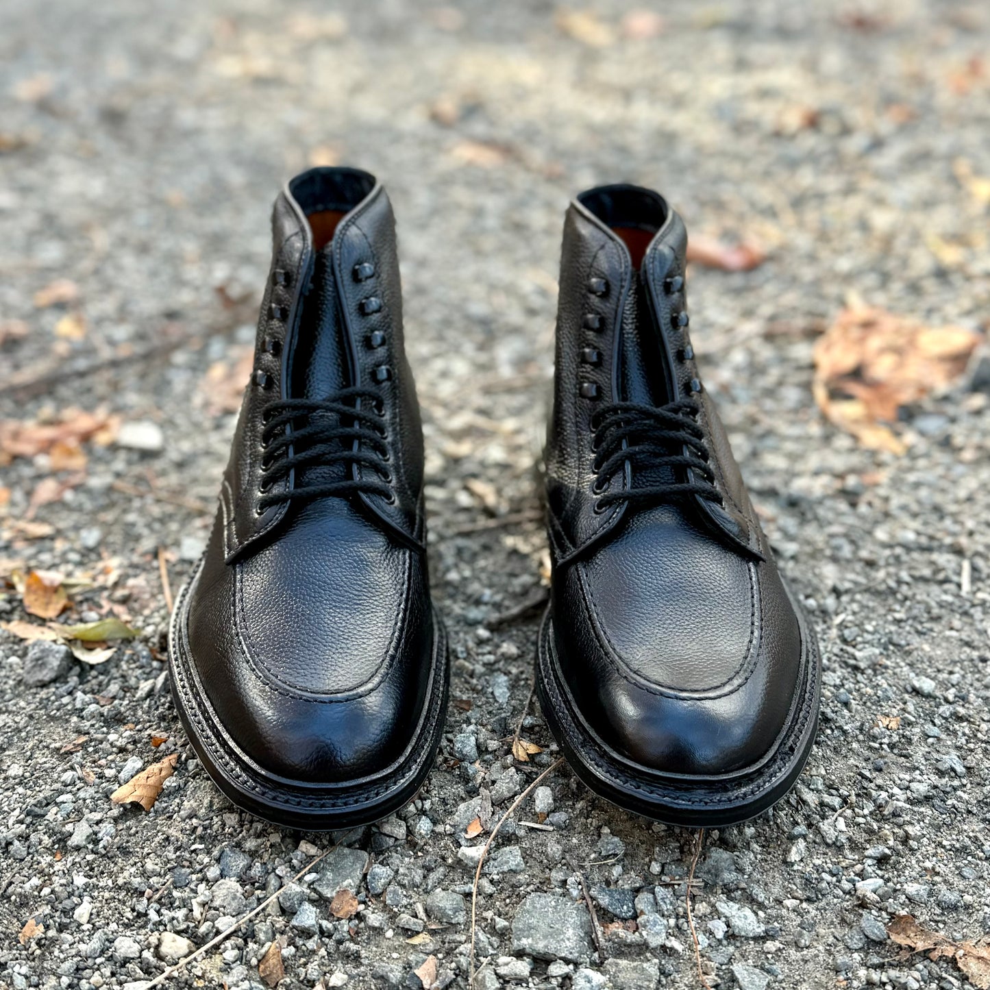 A pair of Alden Madison D6956LH "Han" Boots in Black JS Grain with eyelets, crafted on the Barrie Last, placed side by side on a gravel surface with scattered leaves.