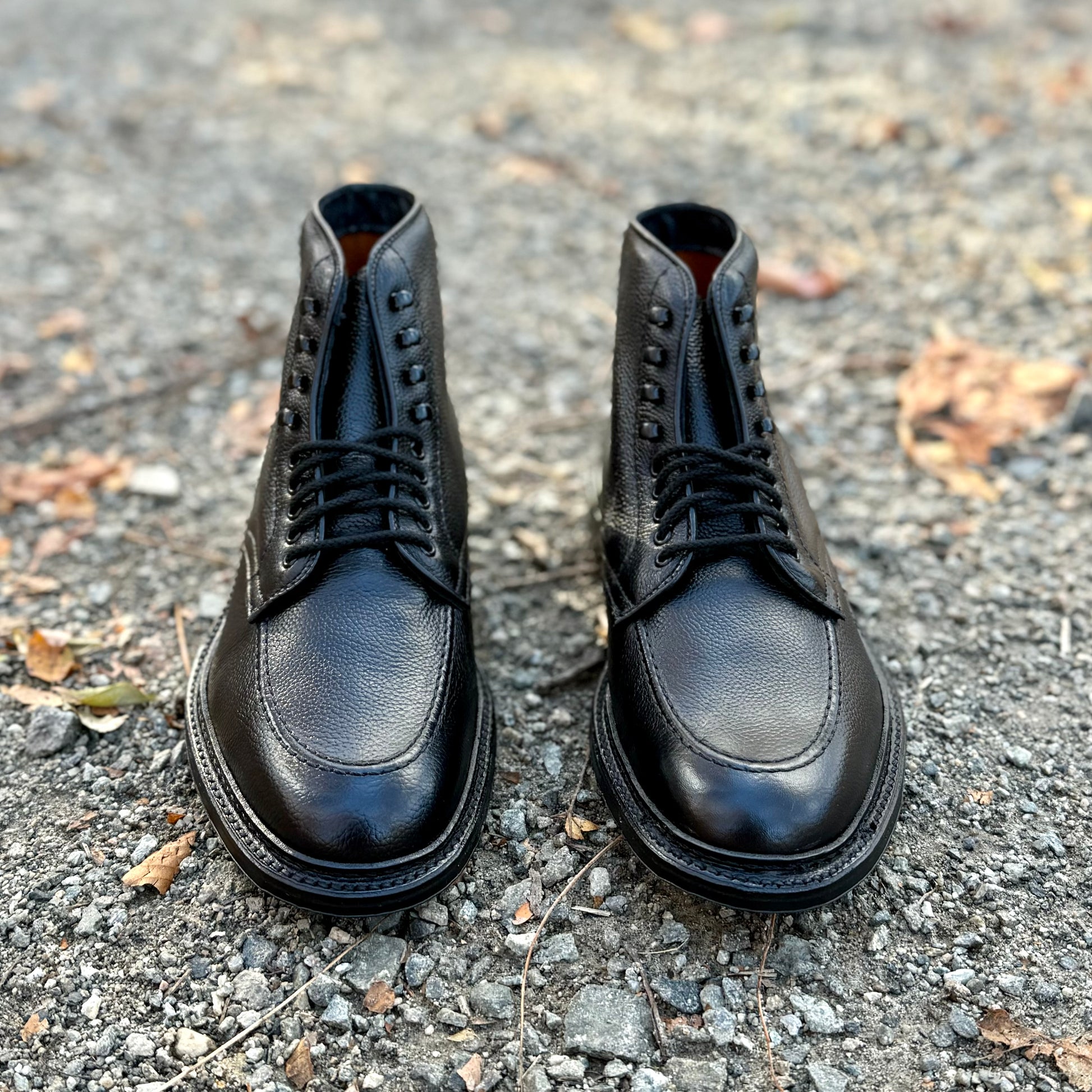 A pair of Alden Madison D6956LH "Han" Boots in Black JS Grain with eyelets, crafted on the Barrie Last, placed side by side on a gravel surface with scattered leaves.