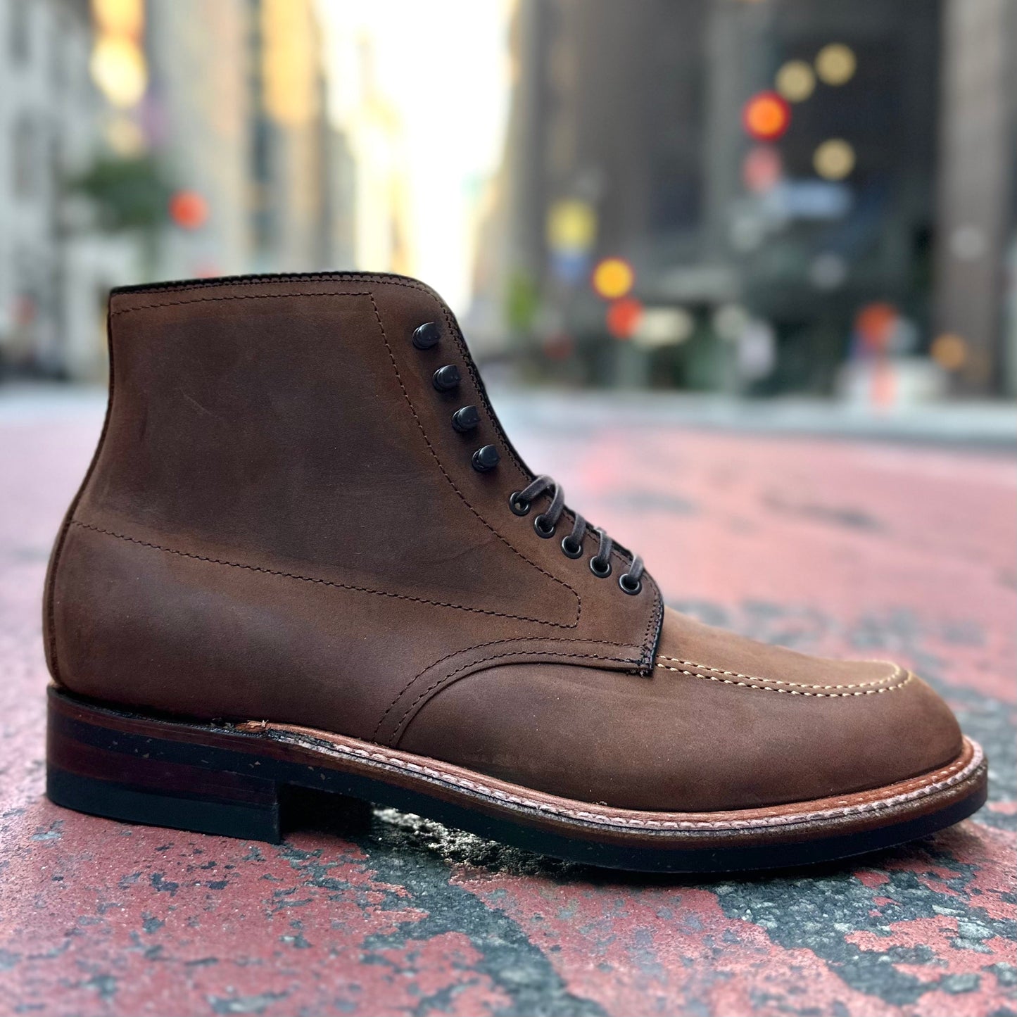 Set against a textured street, Alden Madisons D2909H Indy Boot in Tobacco Smooth Chamois, featuring black laces and a rugged sole, stands out. The blurred cityscape with buildings and streetlights enhances its urban vibe, while the Trubalance Last design ensures style and comfort for city adventurers.