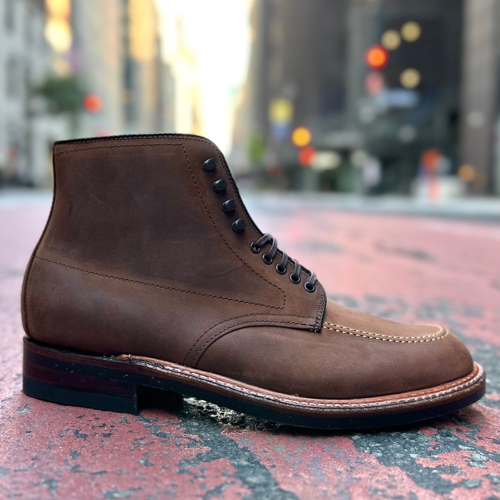 Set against a textured street, Alden Madisons D2909H Indy Boot in Tobacco Smooth Chamois, featuring black laces and a rugged sole, stands out. The blurred cityscape with buildings and streetlights enhances its urban vibe, while the Trubalance Last design ensures style and comfort for city adventurers.