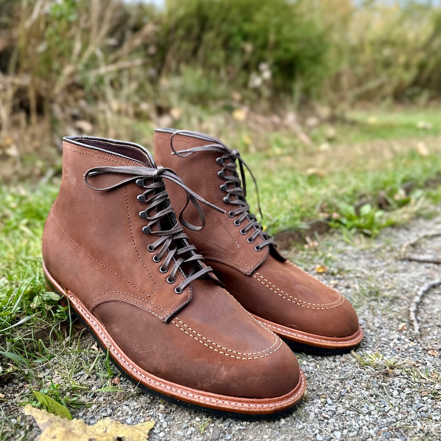The Alden Madison D2909H Indy Boots in Tobacco Smooth Chamois, featuring dark laces and visible stitching, are artfully crafted on the Trubalance Last. They rest on a gravel path surrounded by grass and foliage, exuding a stylish and durable charm.
