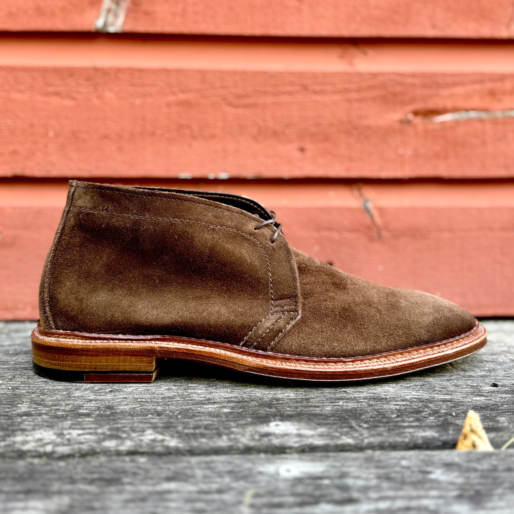The Alden Madison 1492 - Unlined Chukka in Brown Suede, featuring laces and a Single Flex Leather Sole, is displayed on a weathered wooden surface with an orange wooden wall as the backdrop.
