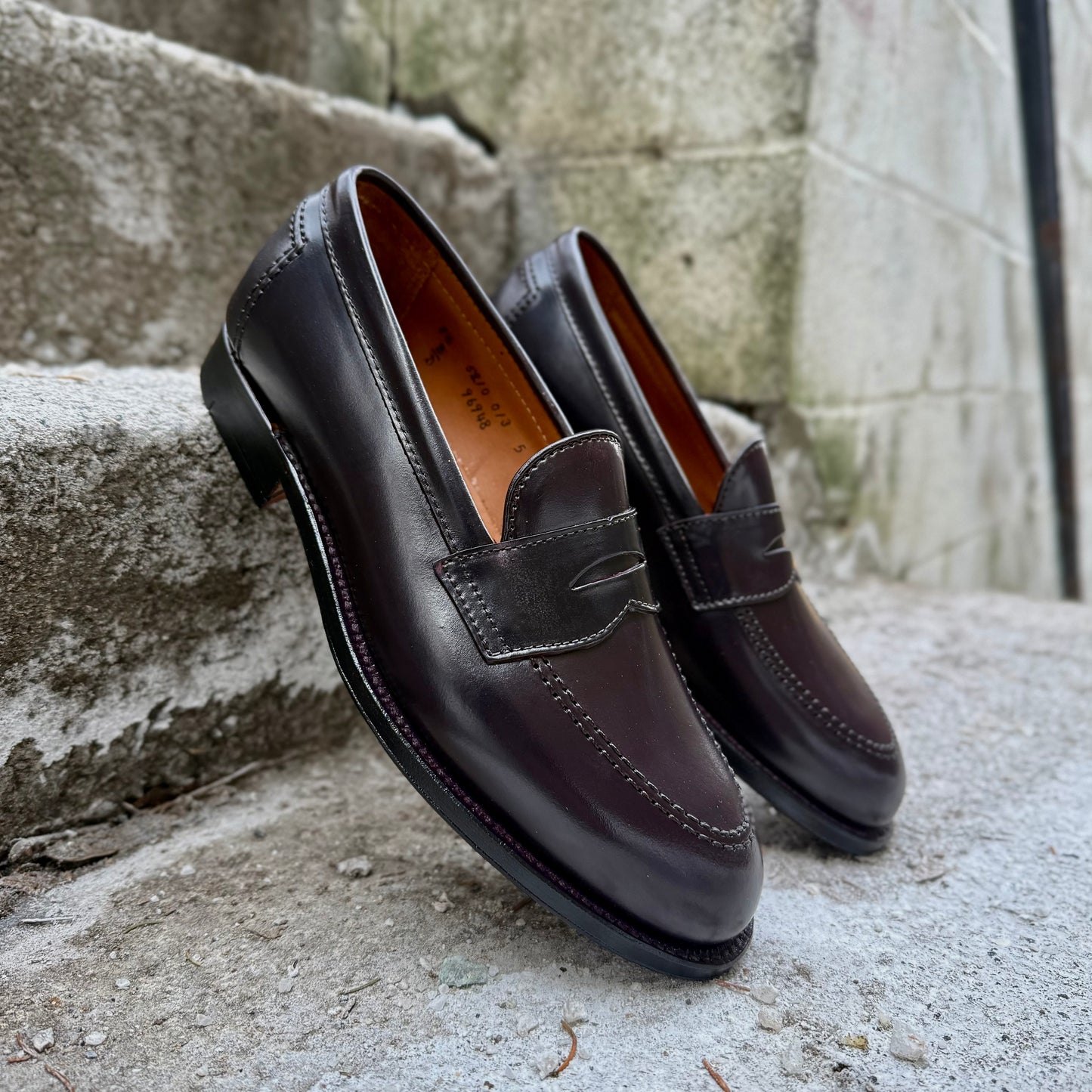 A pair of ALDEN SHOES 96948 - Copley Penny Loafer in Color 8 Shell with visible stitching sits on rough concrete steps near a stone wall, highlighting its classic Copley Last and refined leather sole.