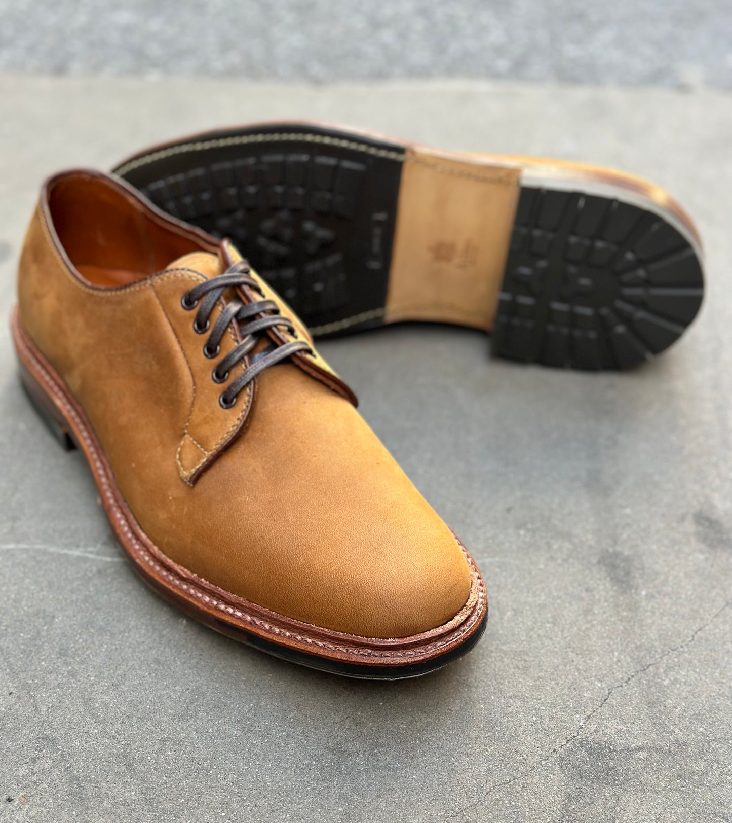 A pair of Alden Madison 9475C - Plain Toe Blucher in Tan Smooth Chamois, one upright showing the upper and laces, the other on its side revealing a black Commando sole, rest on a smooth grey surface.