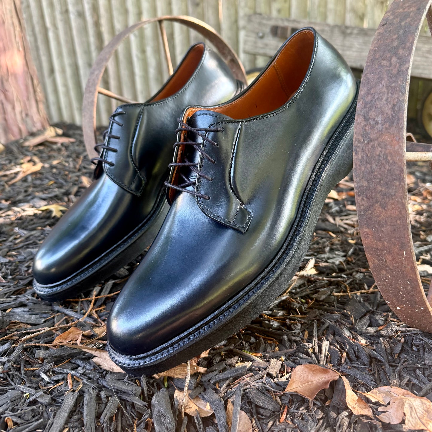 A pair of ALDEN SHOES N1401 Black Calf Plain Toe Blucher with laces are displayed outdoors on dark mulch, resting against a rusty metal wagon wheel with a wooden fence in the background. Crafted from shiny black calfskin leather, these shoes feature a comfortable wedge rubber sole and are built on the classic Barrie Last, blending timeless style with modern durability.