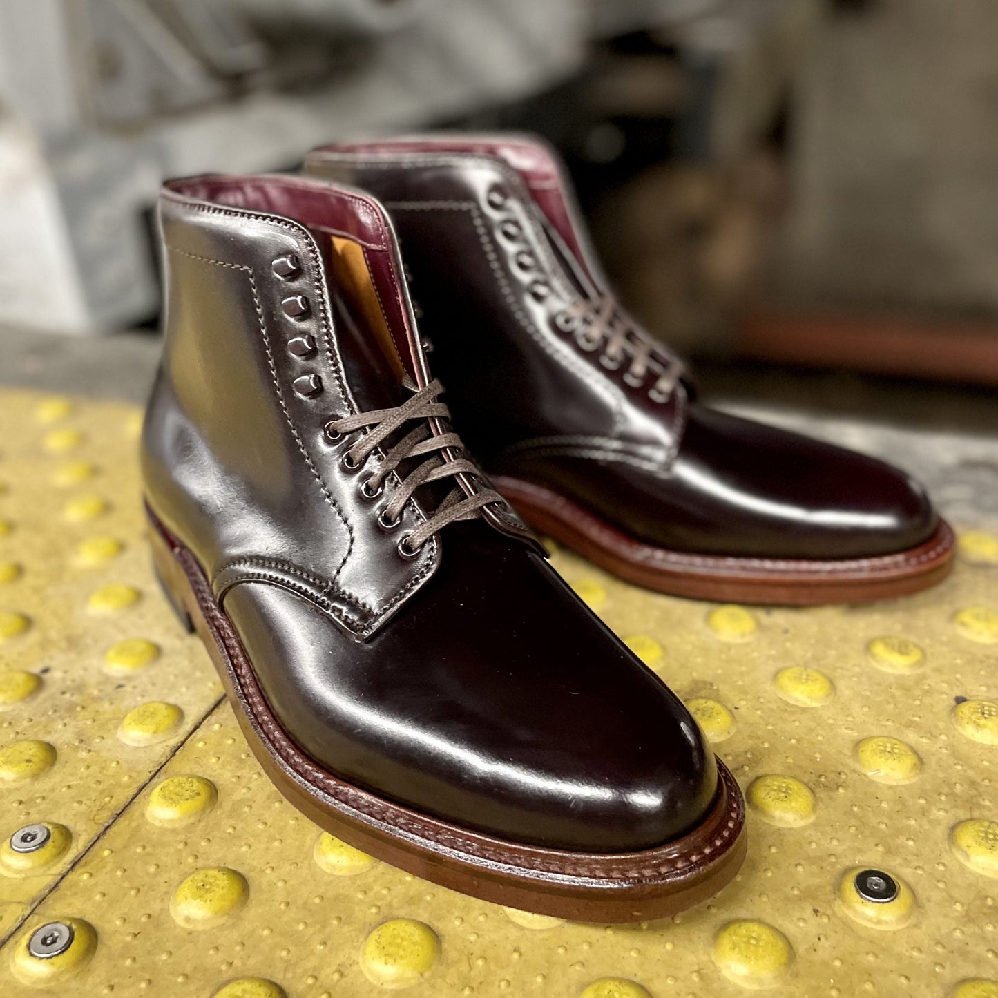 A pair of Alden Madison D1854H - Plain Toe Boot w Neocork Sole in Color 8 Shell, crafted from polished Shell Cordovan leather with laces on the Barrie Last, is placed on a yellow textured surface featuring raised dots.