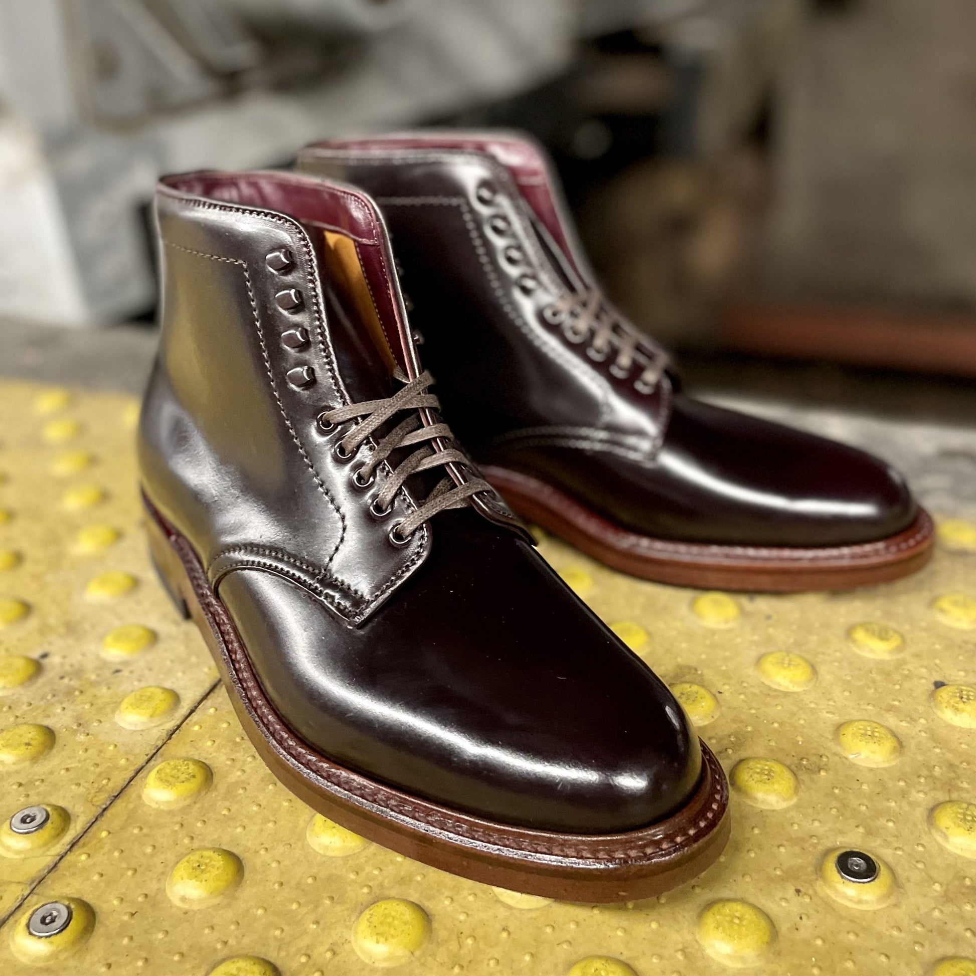 A pair of Alden Madison D1854H - Plain Toe Boot w Neocork Sole in Color 8 Shell, crafted from polished Shell Cordovan leather with laces on the Barrie Last, is placed on a yellow textured surface featuring raised dots.