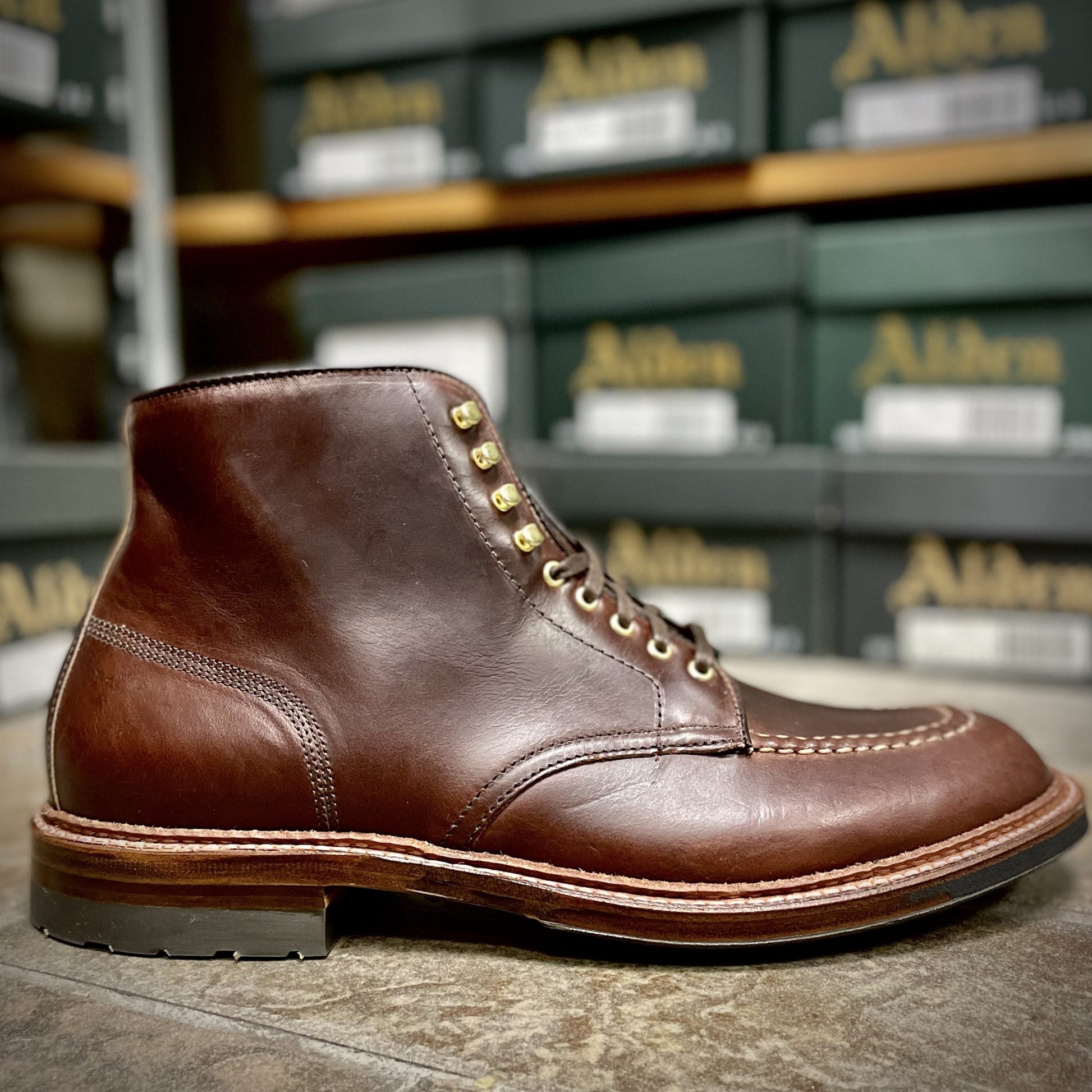A close-up of Alden Madisons D1932HC - Dirty Bones Indy Boot in Brown Chromexcel, crafted with polished leather and featuring a stitched toe and gold eyelets, is showcased against a backdrop of neatly arranged green shoeboxes.