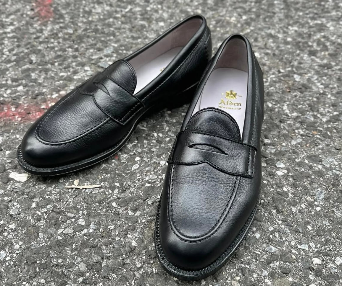 The Alden 9695F Flex Penny Loafer in Black Calfskin, crafted on the Copley Last with a smooth toe and textured upper, is shown on gray asphalt. The Alden logo appears inside one shoe.