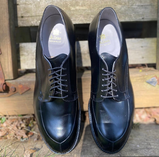 A pair of Alden Madison 539 - Modified V-Tip dress shoes in Black Calfskin with a wide toebox, featuring laces, are placed side by side on a wooden surface outdoors with leaves scattered around.