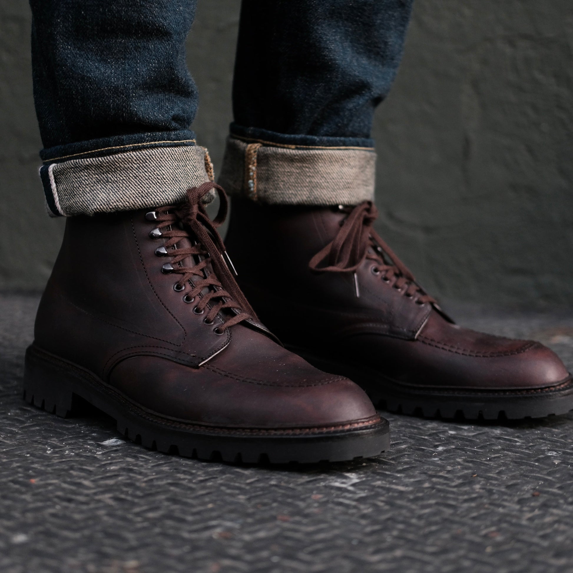 A person is wearing Alden Madisons 404 - Indy Boot in Kudu (Oiled CXL), polished dark brown lace-up boots with a durable Vibram Lug Outsole. Theyre paired with cuffed dark blue jeans and resting on a textured metal surface against a muted green wall.