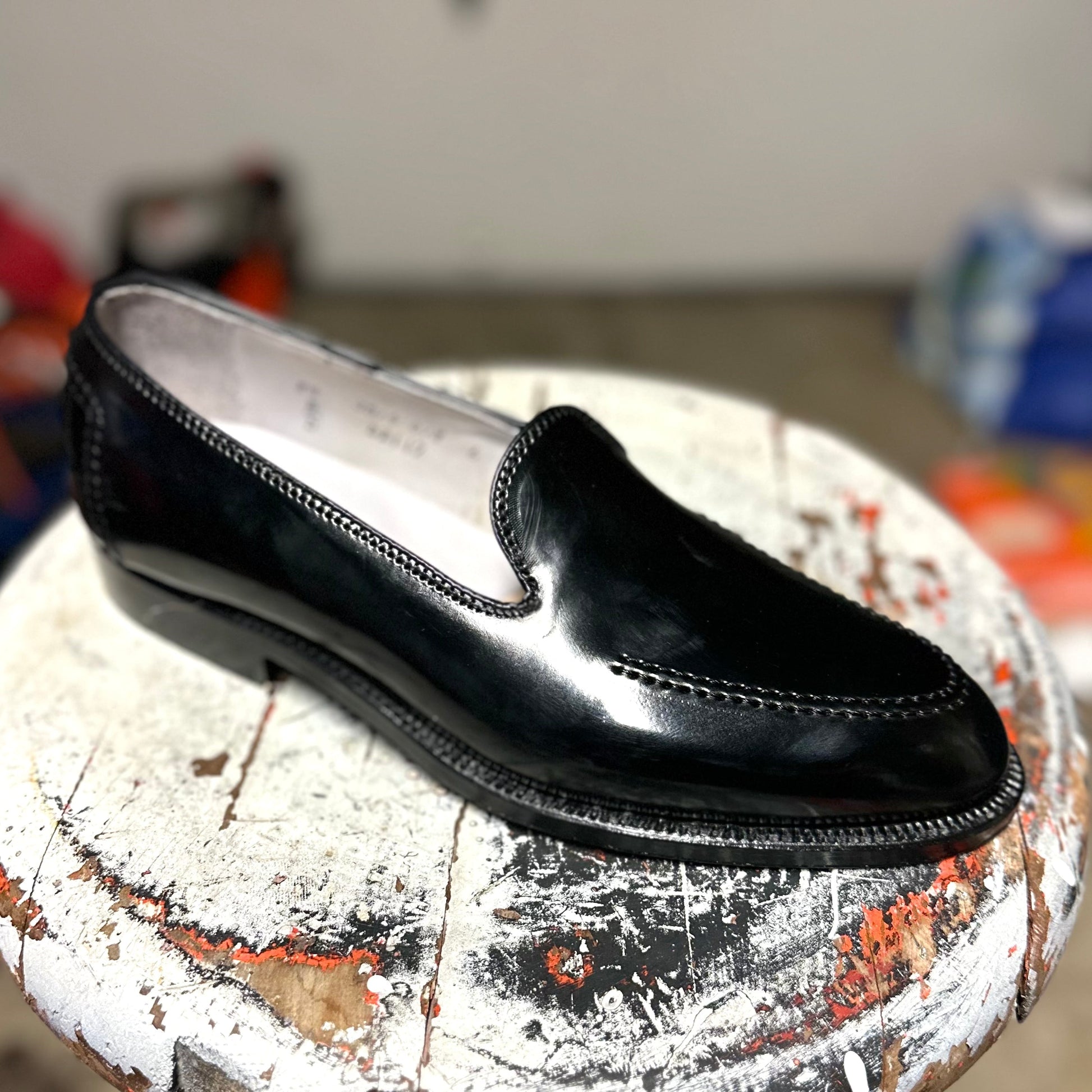 A single, elegant Vanderbilt Slip On in Black Shell Cordovan by Alden Madison with stitching details and a classic Brooks heel rests on a worn, painted wooden stool. The blurred background emphasizes the loafers sophistication.
