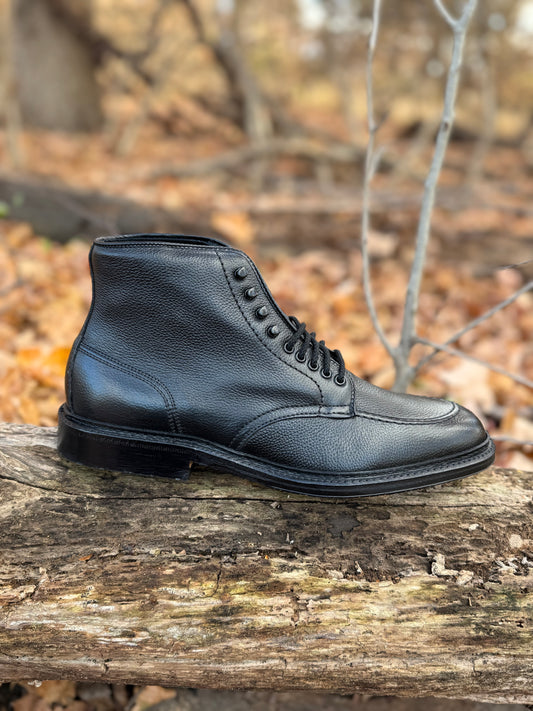 The Alden Madison D6956LH "Han" Boot in Black JS Grain, featuring a textured surface and classic Barrie Last, sits on a fallen log amid brown leaves and bare branches in a forest.