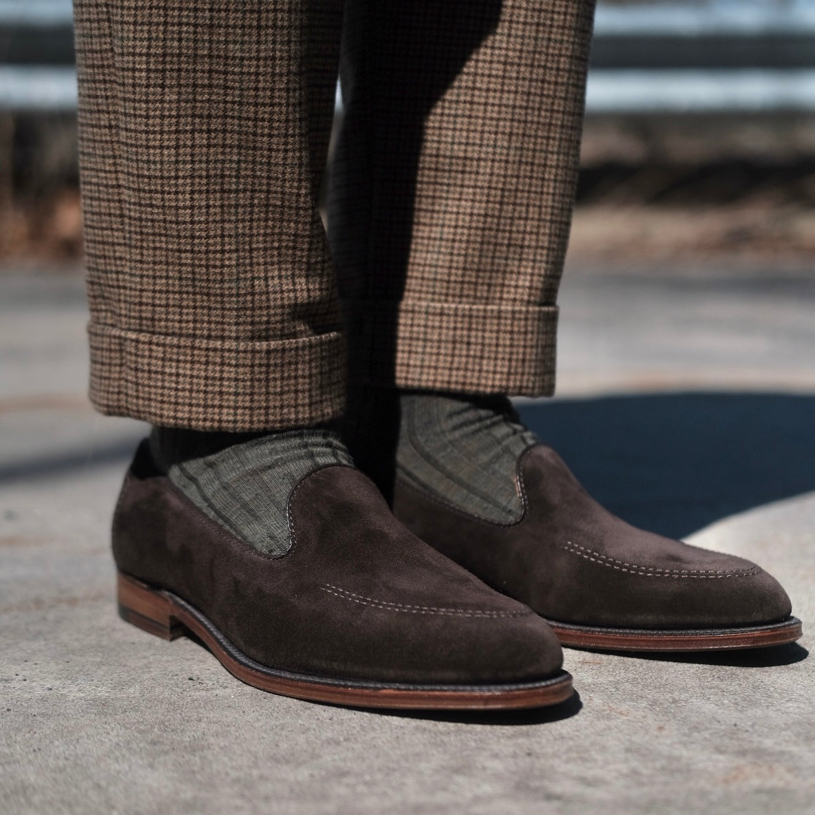 Close-up of a person wearing ALDEN SHOES D5105 - Vanderbilt in Dark Brown Suede with grayish-green patterned socks and brown plaid cuffed trousers, standing on a concrete surface outdoors.