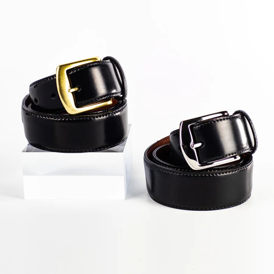 Two Alden Madison MB 5915 - Alden 38mm Black Shell Cordovan Belts, made in the USA, are shown rolled on a white background—one with a gold buckle (elevated on a clear block) and one with a silver buckle.