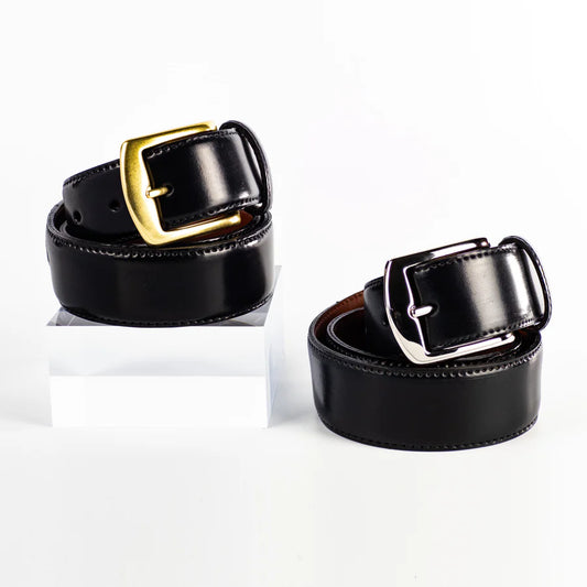 Two Alden Madison MB 5915 - Alden 38mm Black Shell Cordovan Belts, made in the USA, are shown rolled on a white background—one with a gold buckle (elevated on a clear block) and one with a silver buckle.