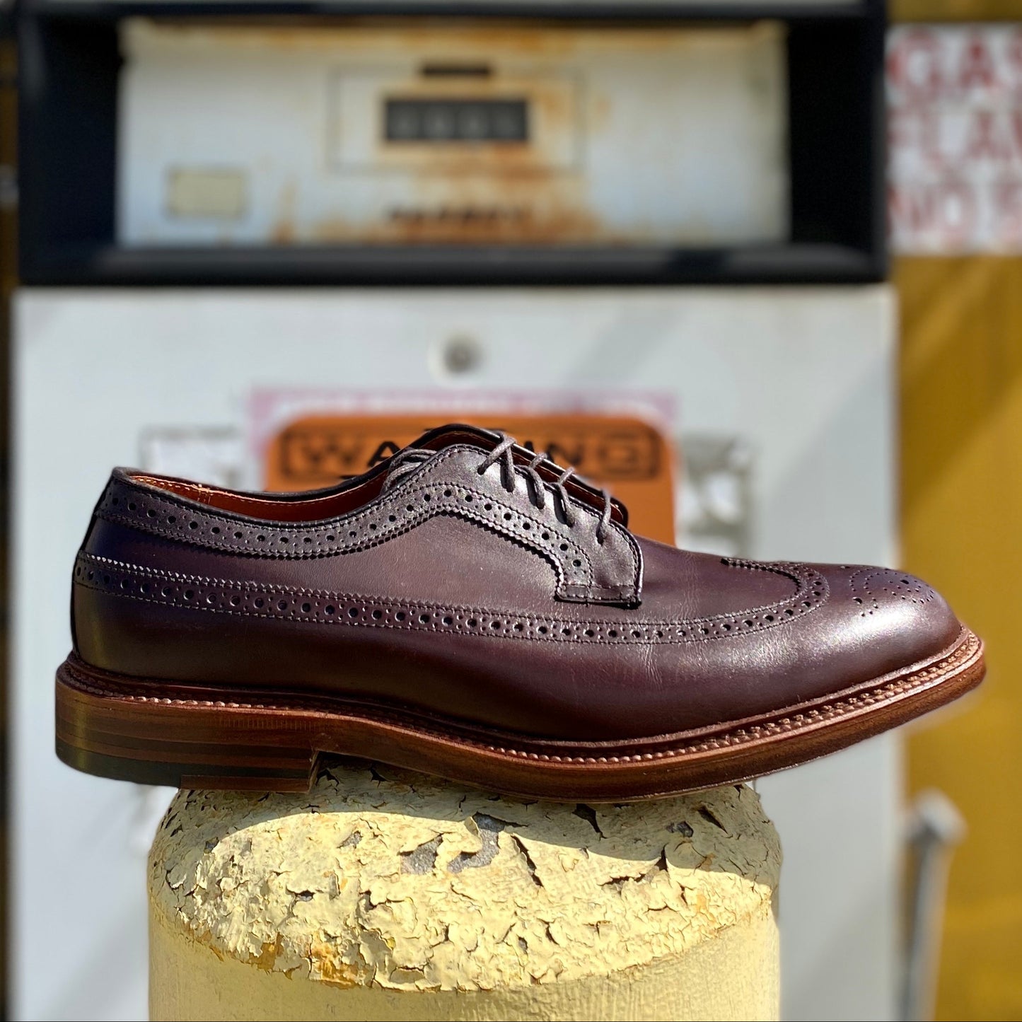 The Alden Madison 976 Long Wing Blucher in polished brown calfskin, featuring brogue detailing and a double waterlock sole, is displayed on a concrete pillar. An old-fashioned UNLEADED gas pump stands under a partly cloudy sky in the background.