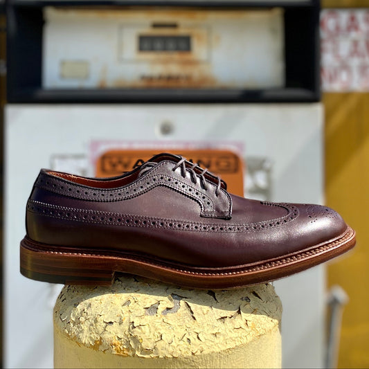 The Alden Madison 976 Long Wing Blucher in polished brown calfskin, featuring brogue detailing and a double waterlock sole, is displayed on a concrete pillar. An old-fashioned UNLEADED gas pump stands under a partly cloudy sky in the background.