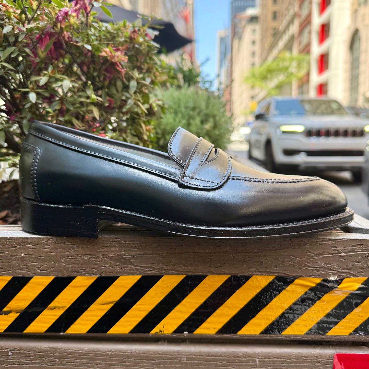 The ALDEN SHOES 5751 - High Vamp Loafer in Black Shell sits on a wood ledge with caution stripes. In the backdrop are plants, city buildings, and a white SUV, highlighting its timeless sophistication crafted on the Aberdeen Last.