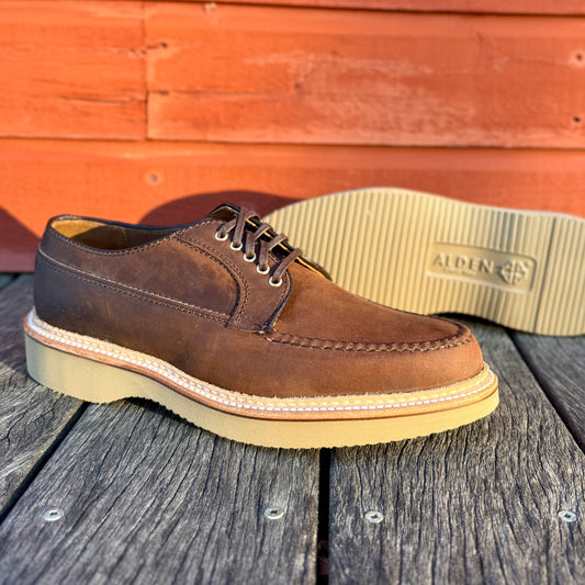 The ALDEN SHOES 73980 - Tobacco Chamois Ranger Mocc with Wedge, featuring white stitching and an ALDEN-marked wedge sole, is displayed on wooden boards against a red wooden wall, with one shoe upright and the other showing the sole.