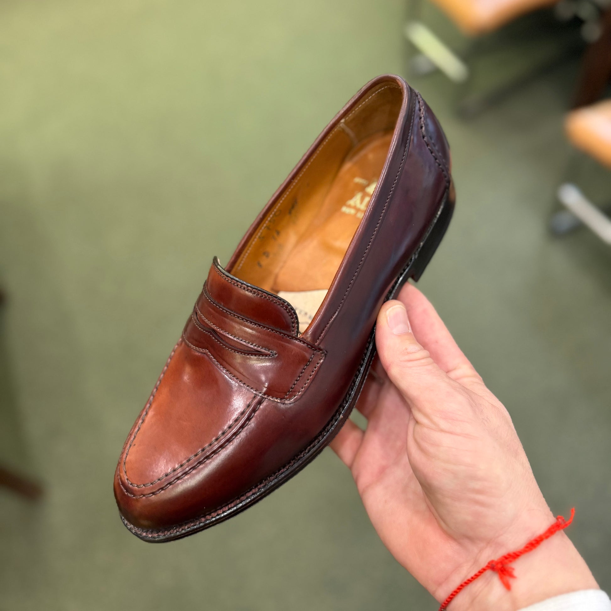 A hand holds the ALDEN SHOES 5750 - Higher Vamp "Midtown" Slip On in Color 8 above a green carpeted floor, with blurred chairs in the background.
