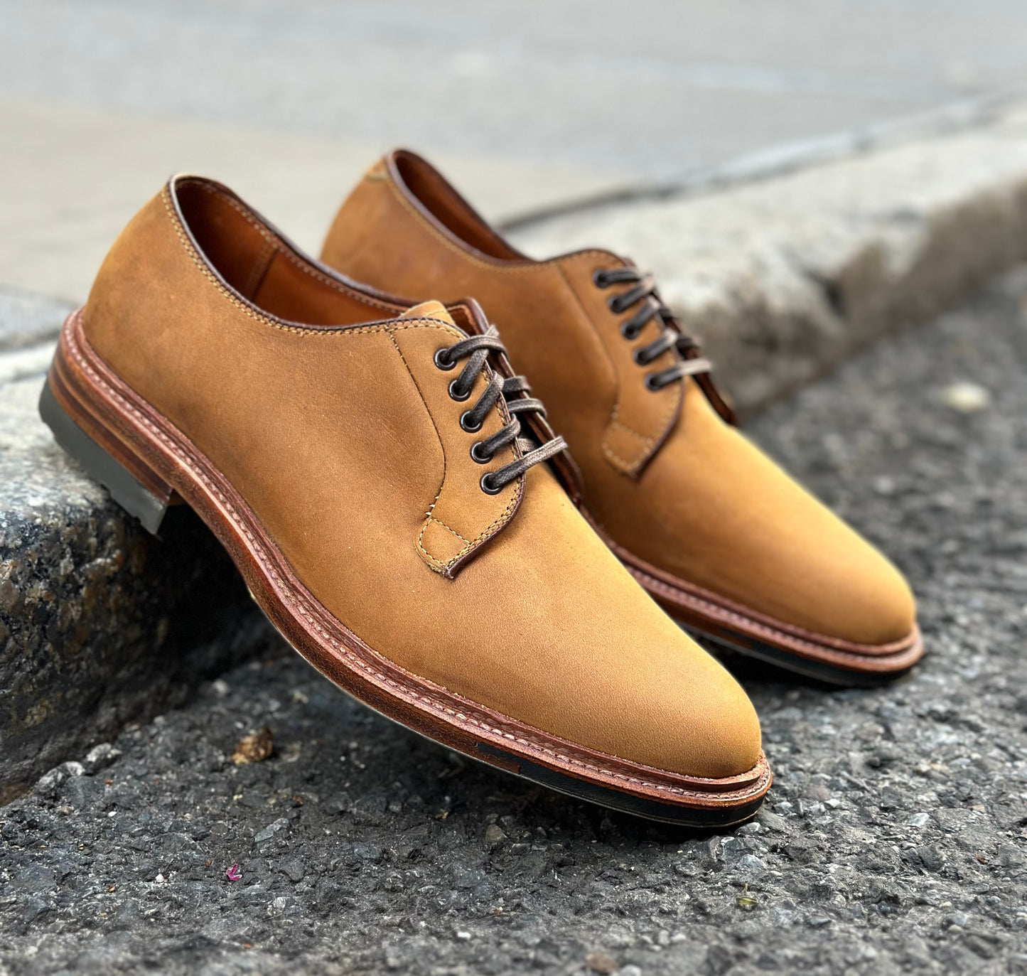 The Alden Madison 9475C - Plain Toe Blucher in Tan Smooth Chamois, featuring dark brown laces and visible stitching along the Commando Sole, sits on textured pavement near a concrete curb.