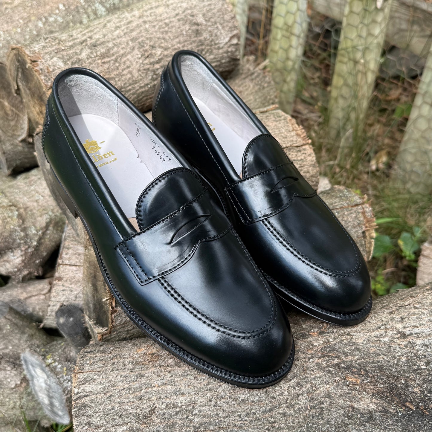 A pair of Alden Madison 96949 - Copley Penny Loafers in Black Shell Cordovan, featuring a classic 270 Flat Welt and crafted on the refined Copley Last, are displayed on stacked logs outdoors, with wooden fence posts and lush greenery in the background.
