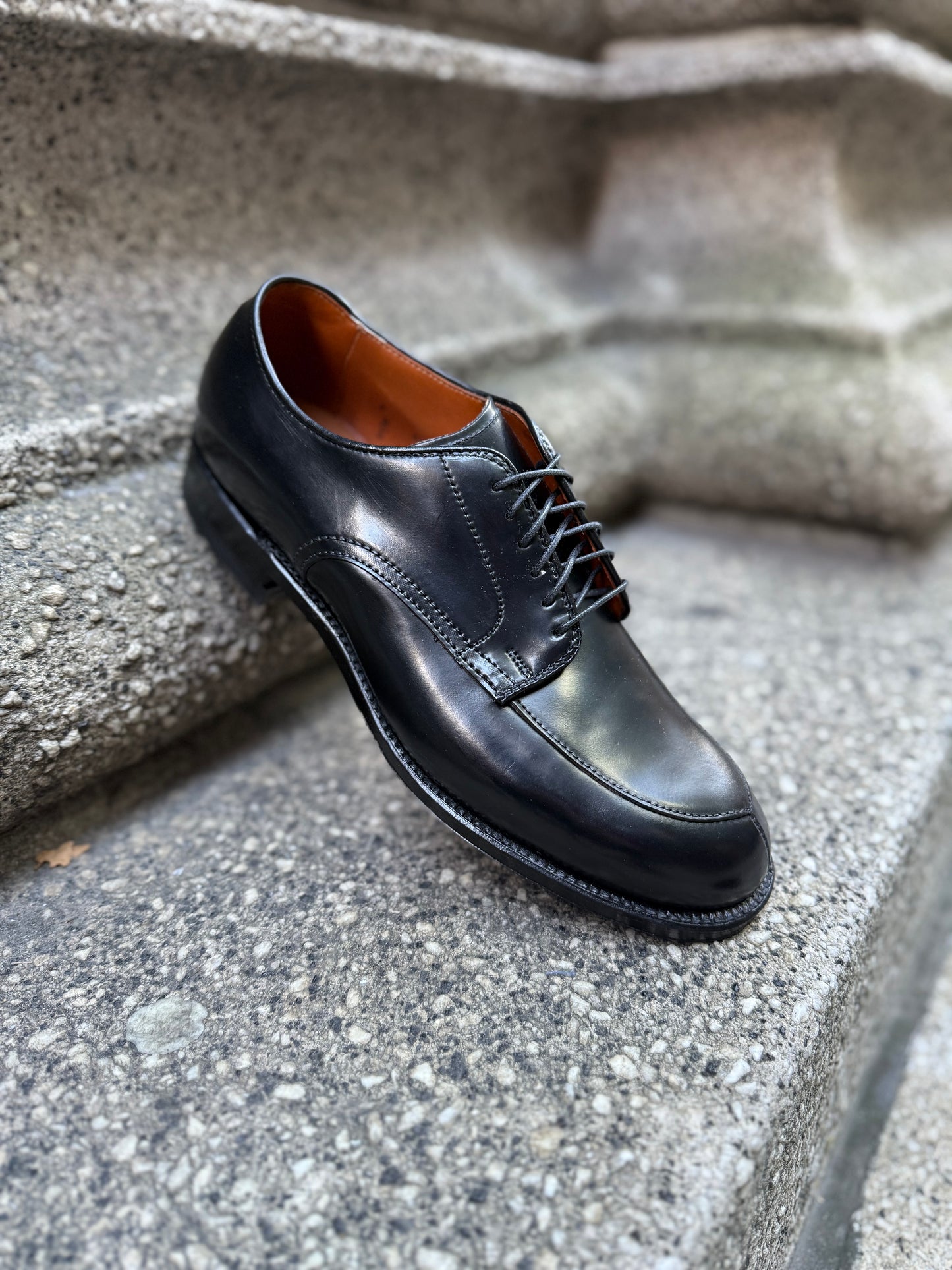 An Alden Madison N6406 - Black Shell V-Tip on 379X dress shoe with laces rests angled on textured stone steps, showcasing polished Shell Cordovan leather, a brown inner lining, and a durable rubber heel.