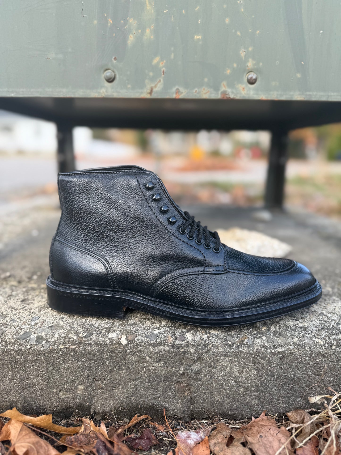 The Alden Madison D6956LH "Han" Boot in Black JS Grain, built on the Barrie Last with textured detailing, rests on outdoor concrete among autumn leaves, its rugged elegance highlighted by a softly blurred background.