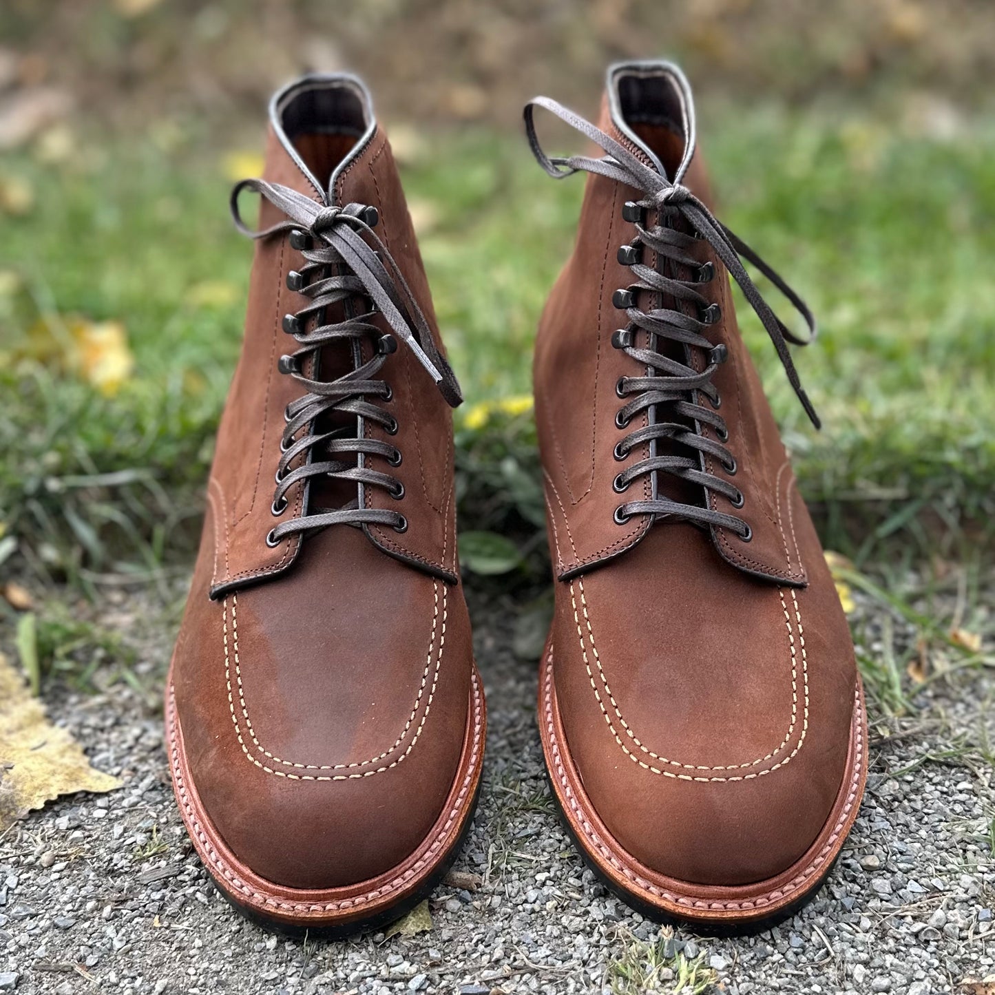 A pair of Alden Madisons D2909H - Indy Boot in Tobacco Smooth Chamois, featuring dark laces and detailed stitching on the Trubalance Last, is set on a gravel path with lush green grass as the backdrop.