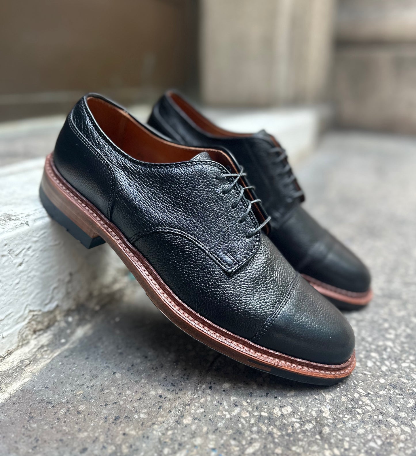 The Alden Madison 22826C - Black JS Grain "Derosa" Blucher dress shoes with brown Commando soles and textured leather uppers are displayed on a concrete surface near a white curb.