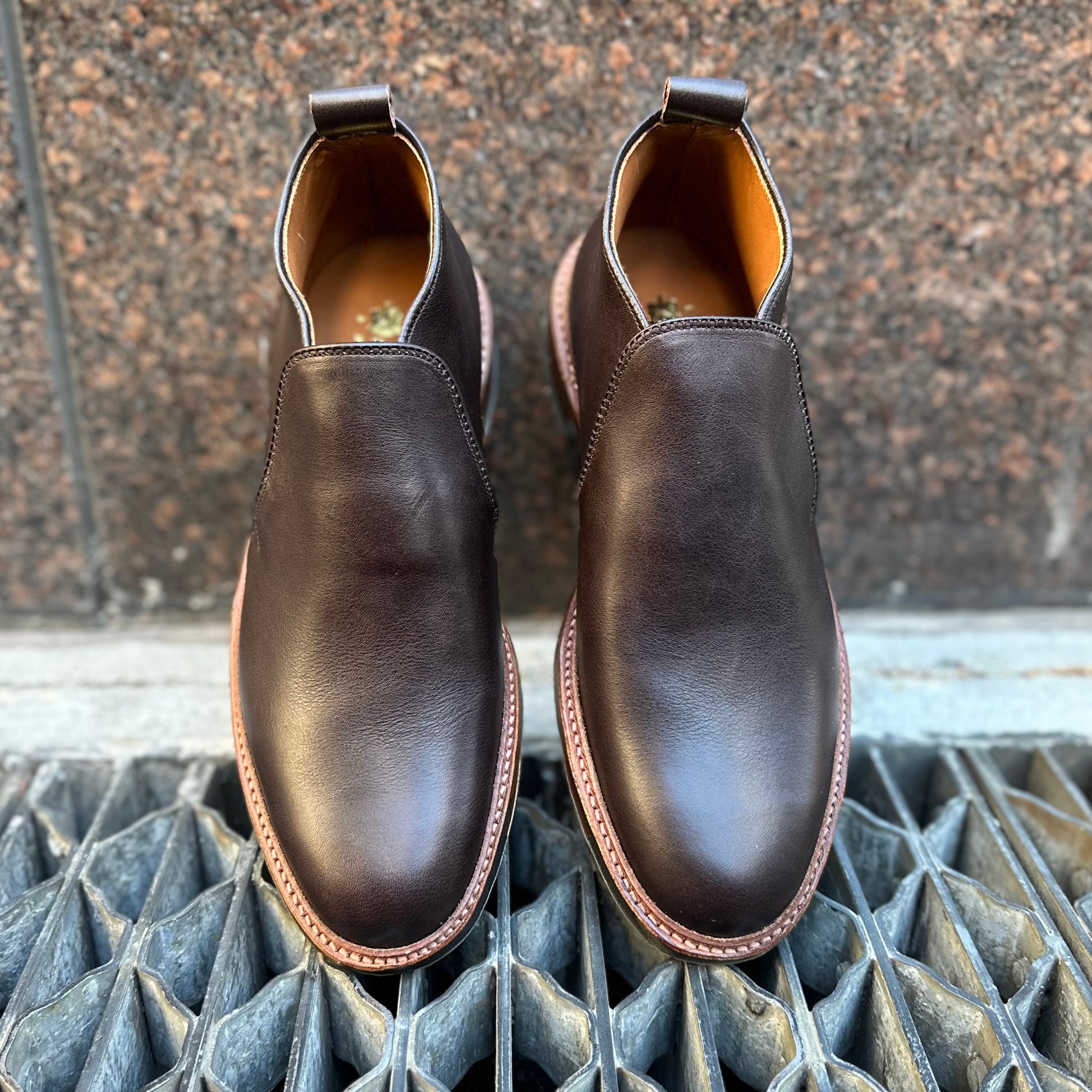 The Alden Madison D2714C - 750 Slip On Chukka in Arabica Lux features dark brown polished leather and light brown Commando Rubber Soles, shown from above on a metal grate with a stone wall background.