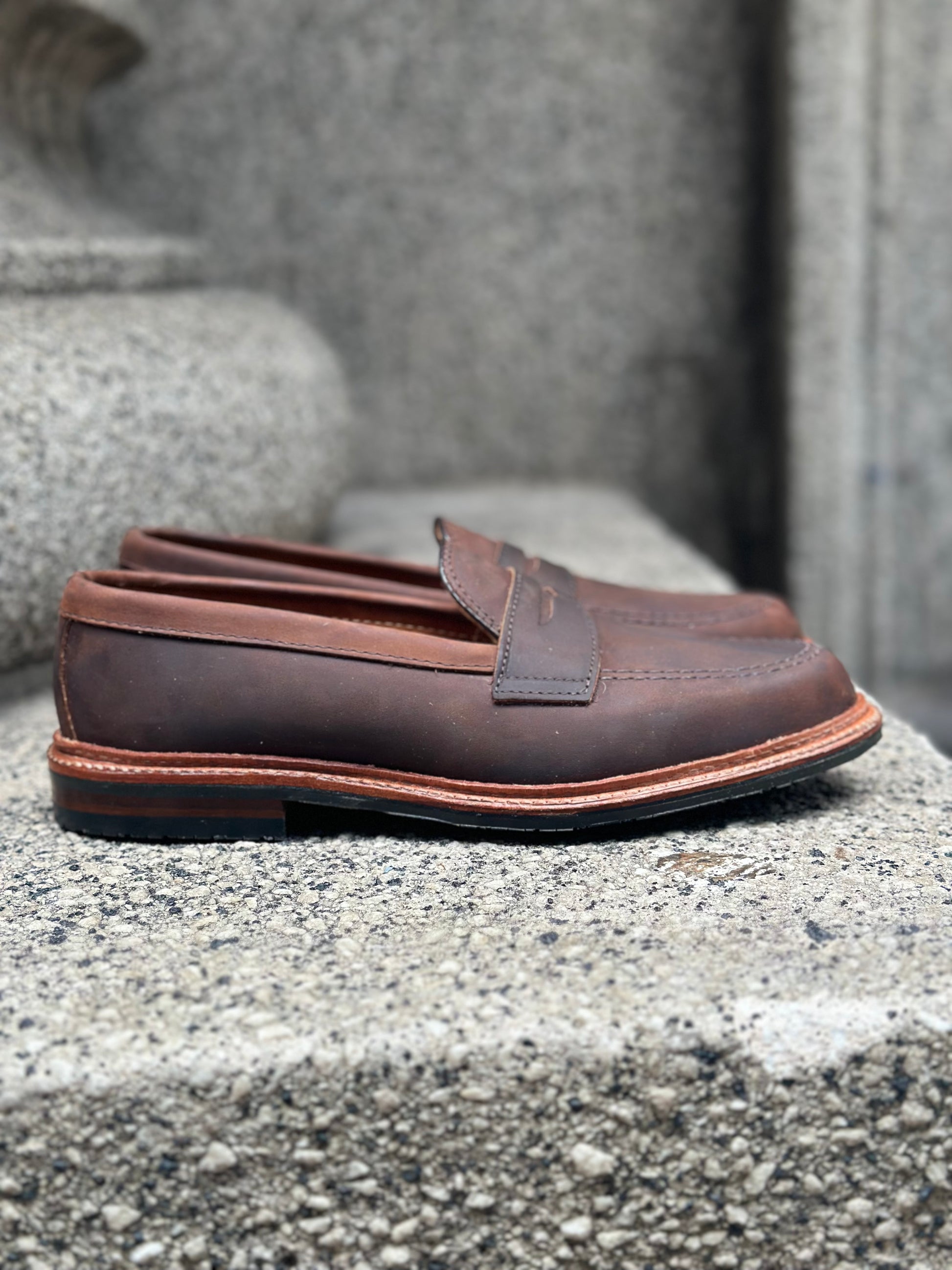 The Alden Madison D2216L - Smooth Tobacco Chamois Penny Loafer, sporting stitched detailing and a durable rubber sole, rests on a textured stone surface with its brown leather finish contrasting against the rough gray background.