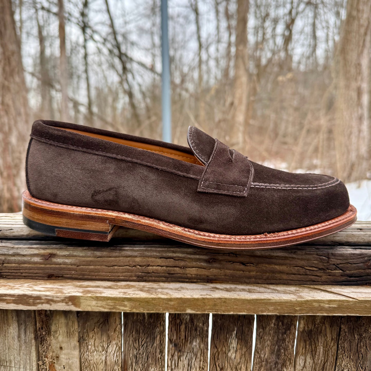 The Alden Madison 6176F Penny Loafer in Dark Chocolate Suede with a flex oiled leather sole sits on a wooden fence outdoors, set against bare trees and a muted forest backdrop.