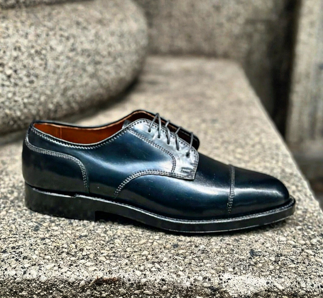 The Alden Madison 2161 - Cap Toe Blucher in Black Shell Cordovan, featuring laces and a double oak leather outsole, is displayed on a textured stone surface.
