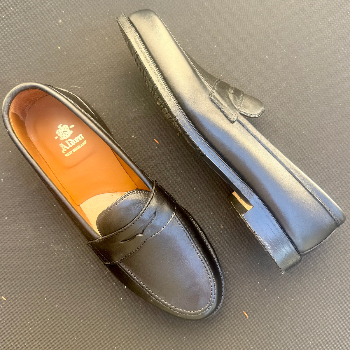 The ALDEN SHOES D5219L Penny Loafer in Black Lady Calf with a rubber sole is displayed, with one shoe laying flat and the other on its side to showcase the durable rubber sole and rich brown interior featuring the Alden logo. Both loafers rest on a plain dark surface.