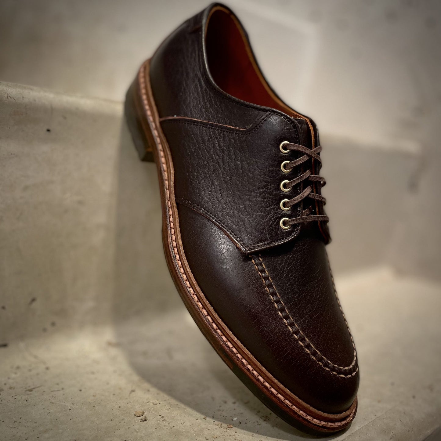 A close-up of Alden Madisons D1613C The Belmont Saddle Blucher Lux in dark brown Arabica calfskin, with light brown stitching and tan laces, crafted on the Barrie Last and displayed on a light-colored textured surface.