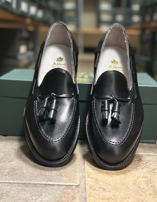 The Alden Madison 660 - Tassel Loafer in Black Calfksin, featuring a leather sole, is shown in front of a green shoebox with blurred shelves of shoes in the background.