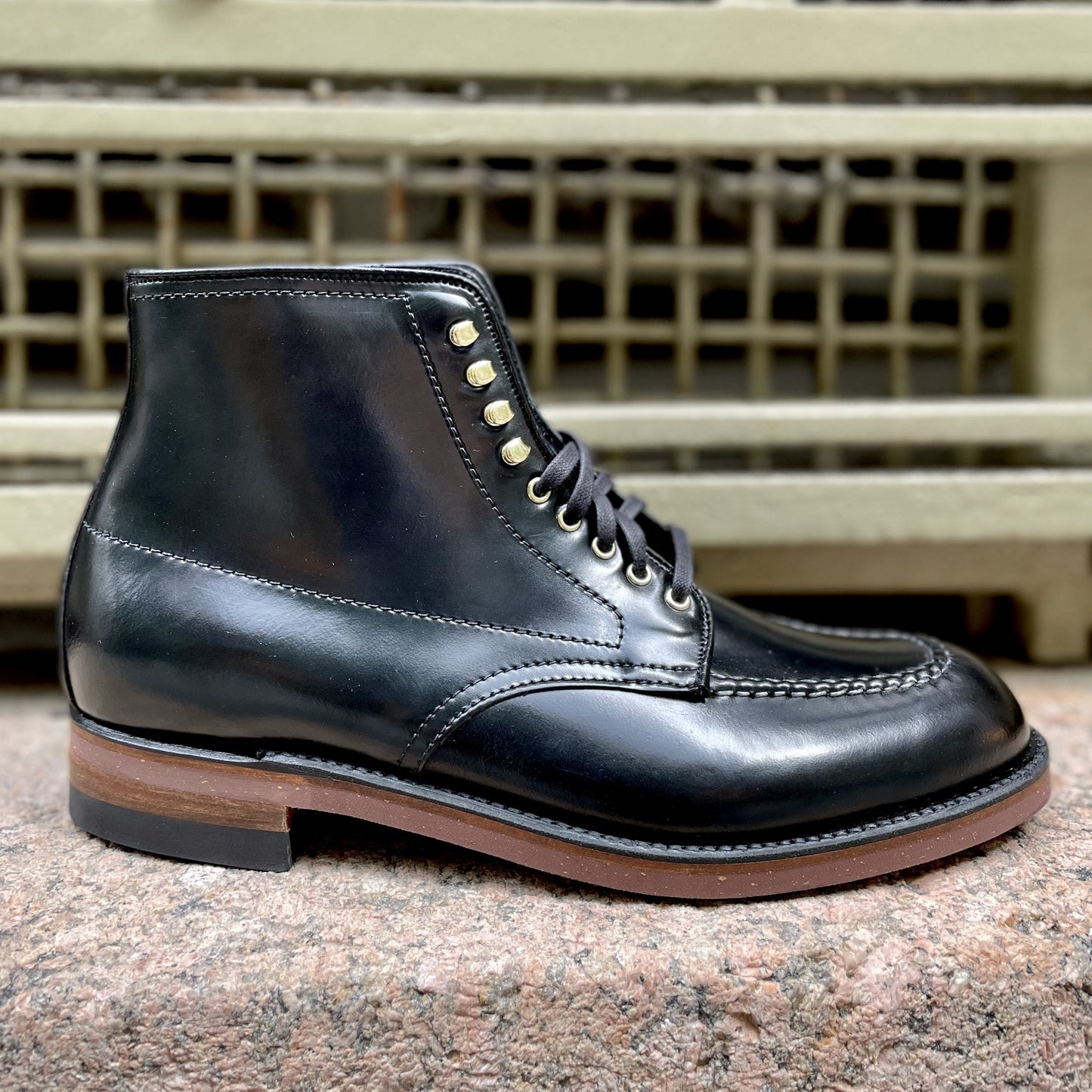 The Alden Madison D1908H Copake Indy Boot in Black Shell, featuring gold eyelets, hand stitching, and a brown sole, is displayed on a stone surface with a metal grate backdrop.