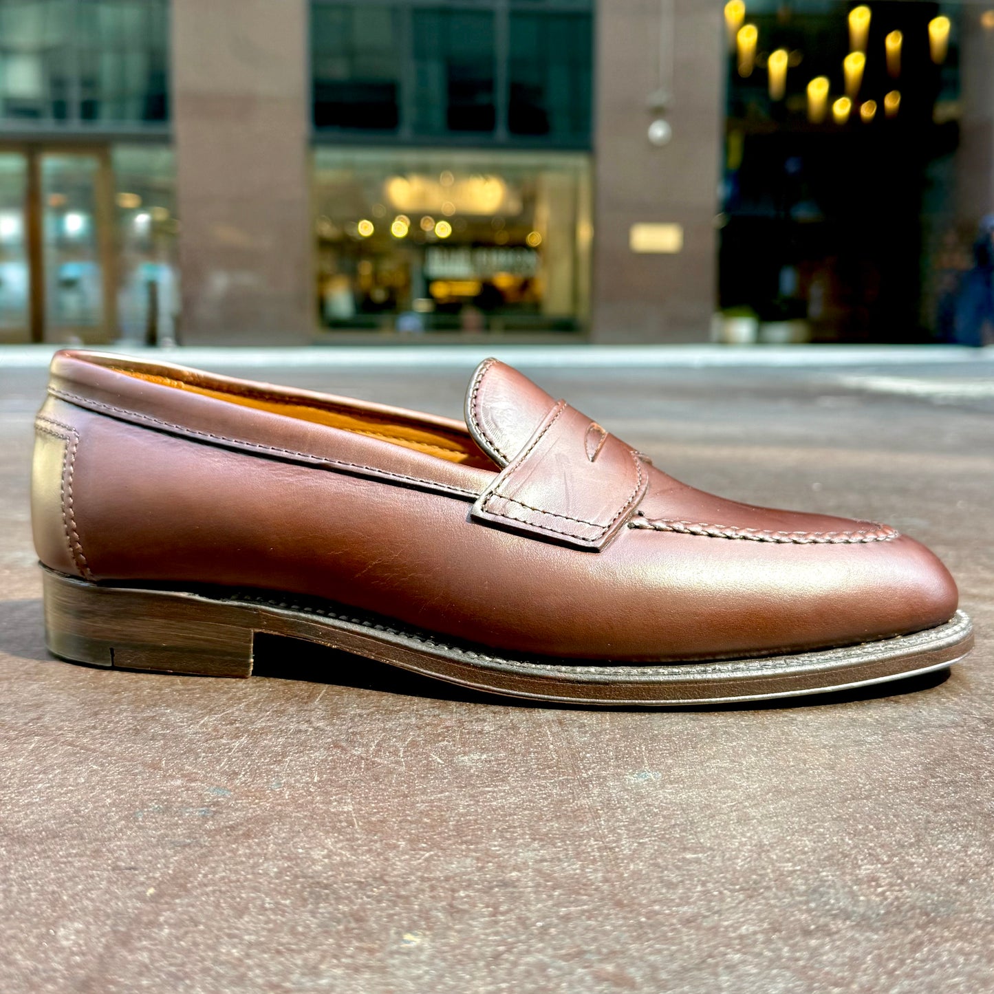 The Alden Madison D5211 - Brown CXL Flex Penny Loafer with a classic leather sole is shown outdoors on a flat surface, with softly blurred city lights and buildings in the background.
